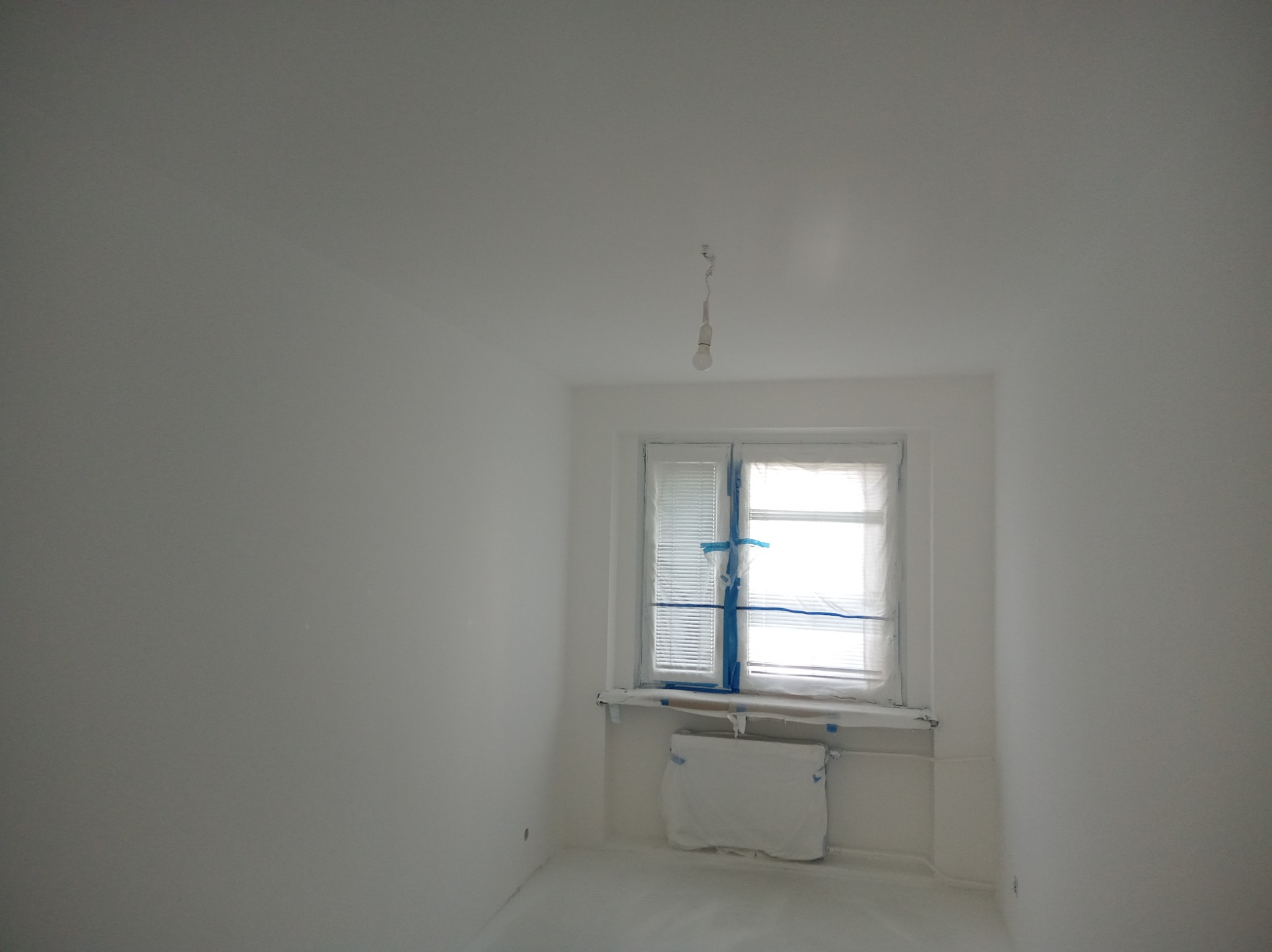 Wnętrze pokoju po malowaniu na biało, z zabezpieczonym oknem i grzejnikiem folią malarską. Widoczna żarówka na kablu. Świeżo pomalowane ściany i podłoga.