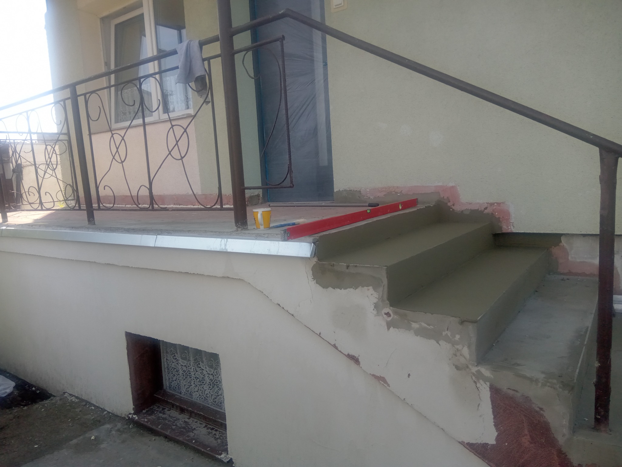Renowacja schodów wejściowych z poziomnicą i kubkiem, świeżo nałożona zaprawa, widoczne ślady wcześniejszych prac, metalowa balustrada i okno w tle.