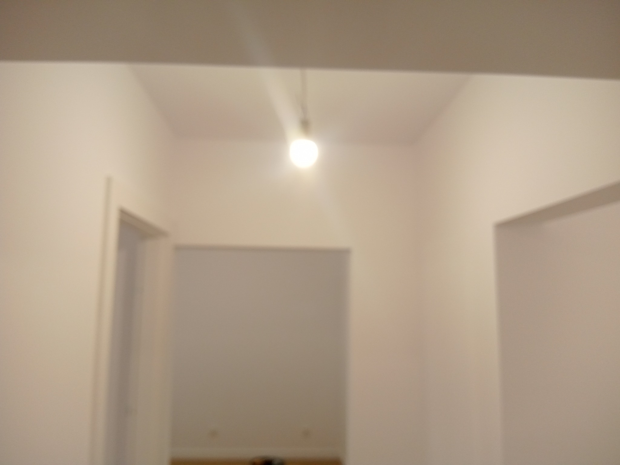 Świeżo pomalowane, jasne ściany wewnątrz pomieszczenia z widocznymi otworami drzwiowymi. Centralnie umieszczona żarówka oświetla wnętrze. Minimalistyczny wystrój.