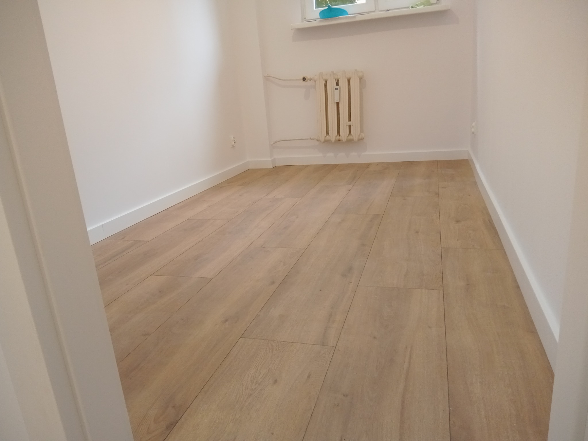 Jasne panele podłogowe w pokoju z białymi ścianami i grzejnikiem. Widok zza framugi drzwi, neutralne oświetlenie, minimalistyczny wystrój.