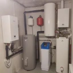 Kompletna instalacja grzewcza w kotłowni, widoczne: pompa ciepła Kaisai, zasobnik Ferroli, zmiękczacz wody z niebieską pokrywą, oraz dwa białe zasobniki Galmet, rury i zawory