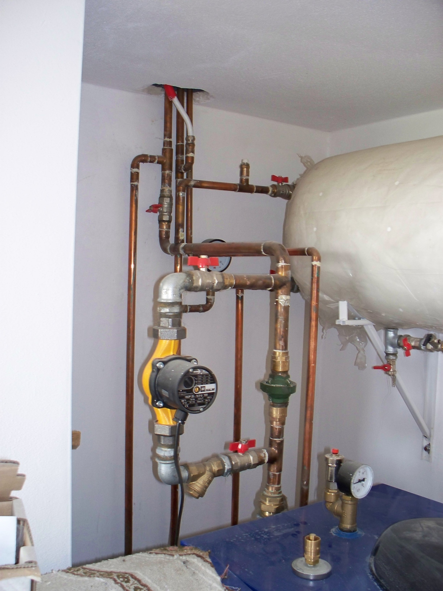 Instalacja centralnego ogrzewania: widok na rury miedziane, pompę obiegową z żółtą obudową i manometr na zbiorniku w pomieszczeniu technicznym.