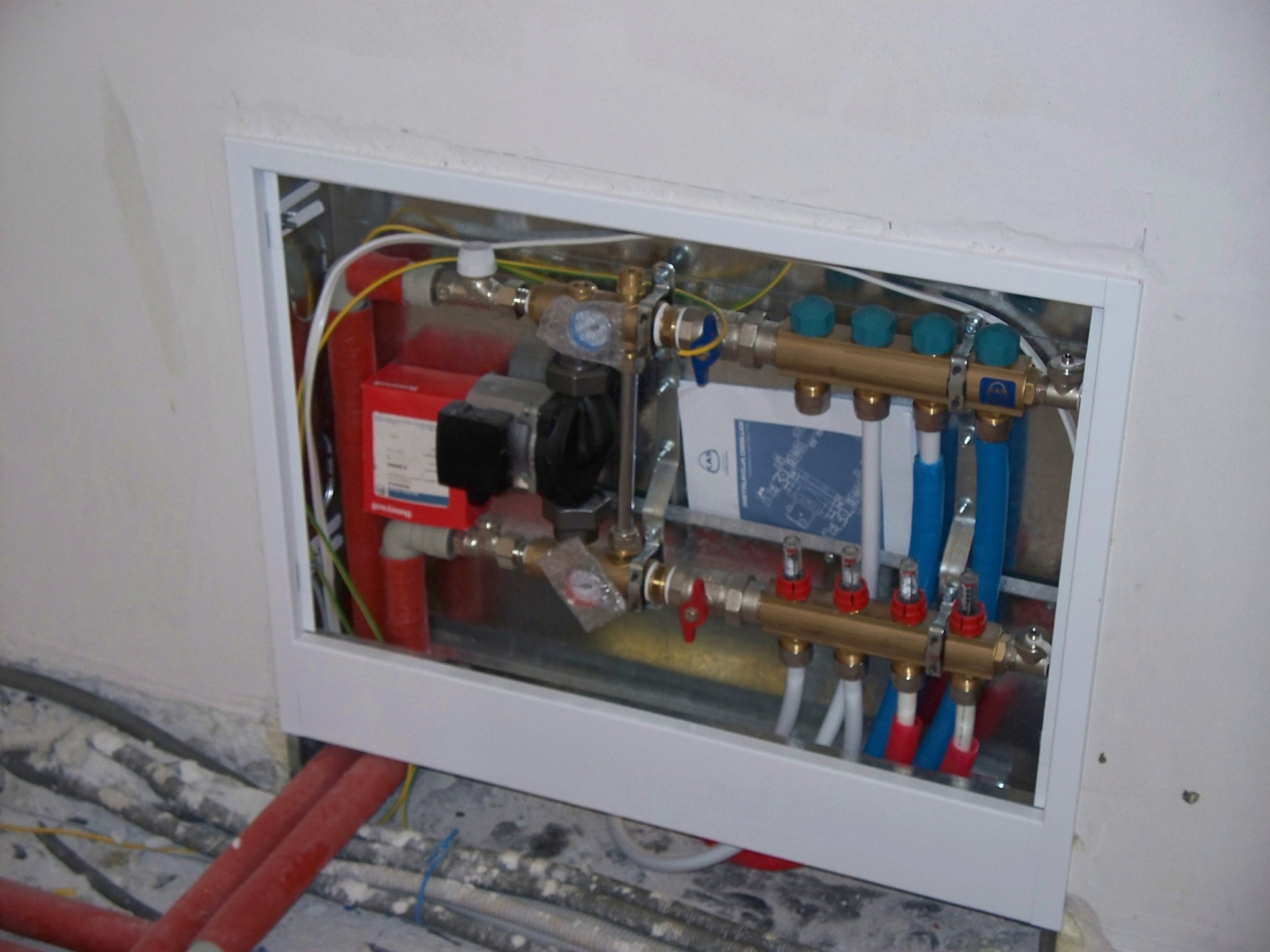 Rozdzielacz ogrzewania podłogowego w metalowej ramie, widoczne rury doprowadzające i odprowadzające wodę, manometry, zawory, pompa obiegowa, instalacja w trakcie montażu.