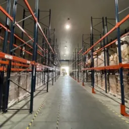 Długi korytarz magazynowy z metalowymi regałami wypełnionymi zapakowanymi kartonami, widok z perspektywy podłogi, z żółto-czarnymi pasami ostrzegawczymi na posadzce.