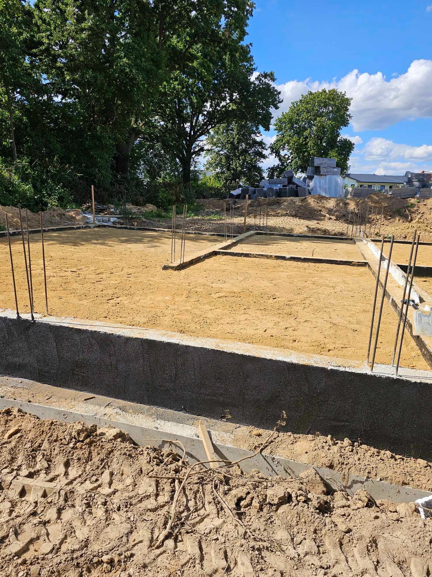 Fundamenty domu w trakcie budowy, widoczne zbrojenia i przygotowane podłoże, w tle drzewa i niebo z chmurami.