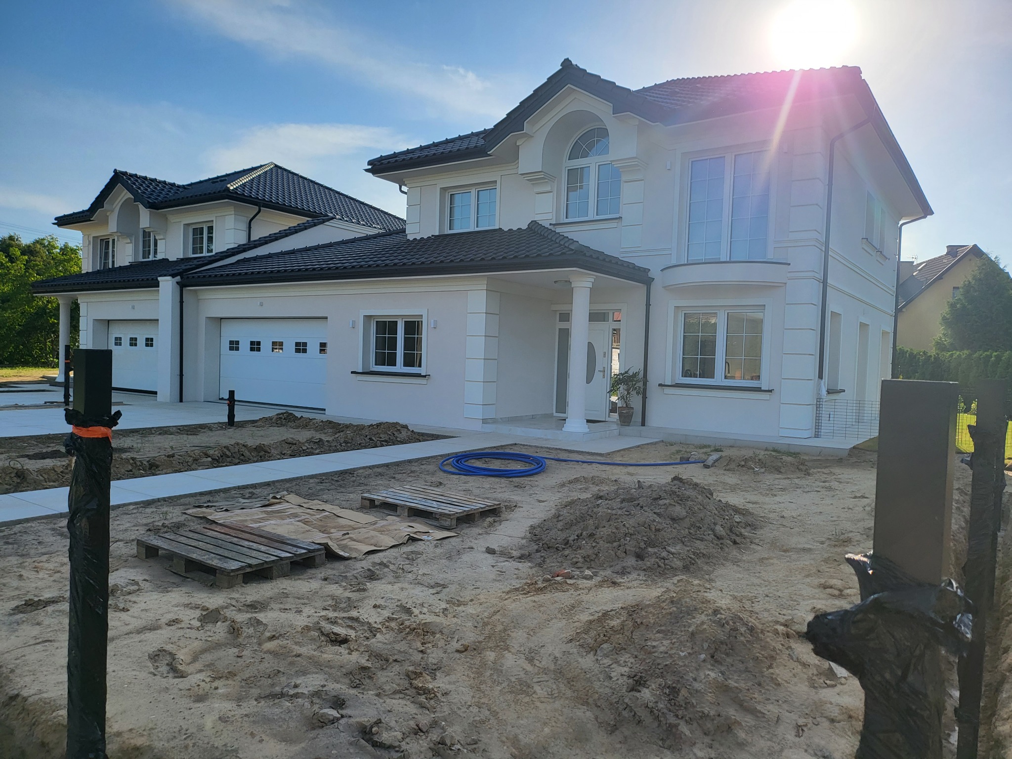 Nowoczesny, dwukondygnacyjny dom z białymi ścianami i ciemną dachówką w trakcie prac wykończeniowych terenu, widoczne palety, rury i świeżo usypana ziemia.