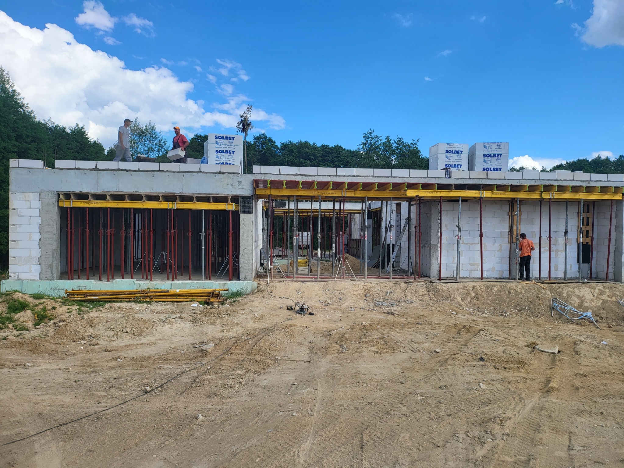 Wznoszenie surowego stanu otwartego domu z bloczków Solbet, widoczne podpory szalunkowe, na górze pracownicy układają kolejne bloczki, ziemisty teren budowy.
