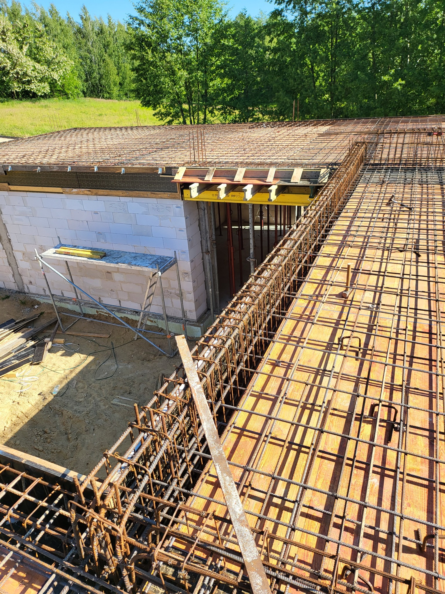 Zbrojenie stropu na deskowaniu, widok z góry, szare bloczki na ścianie w tle, zieleń drzew w oddali.