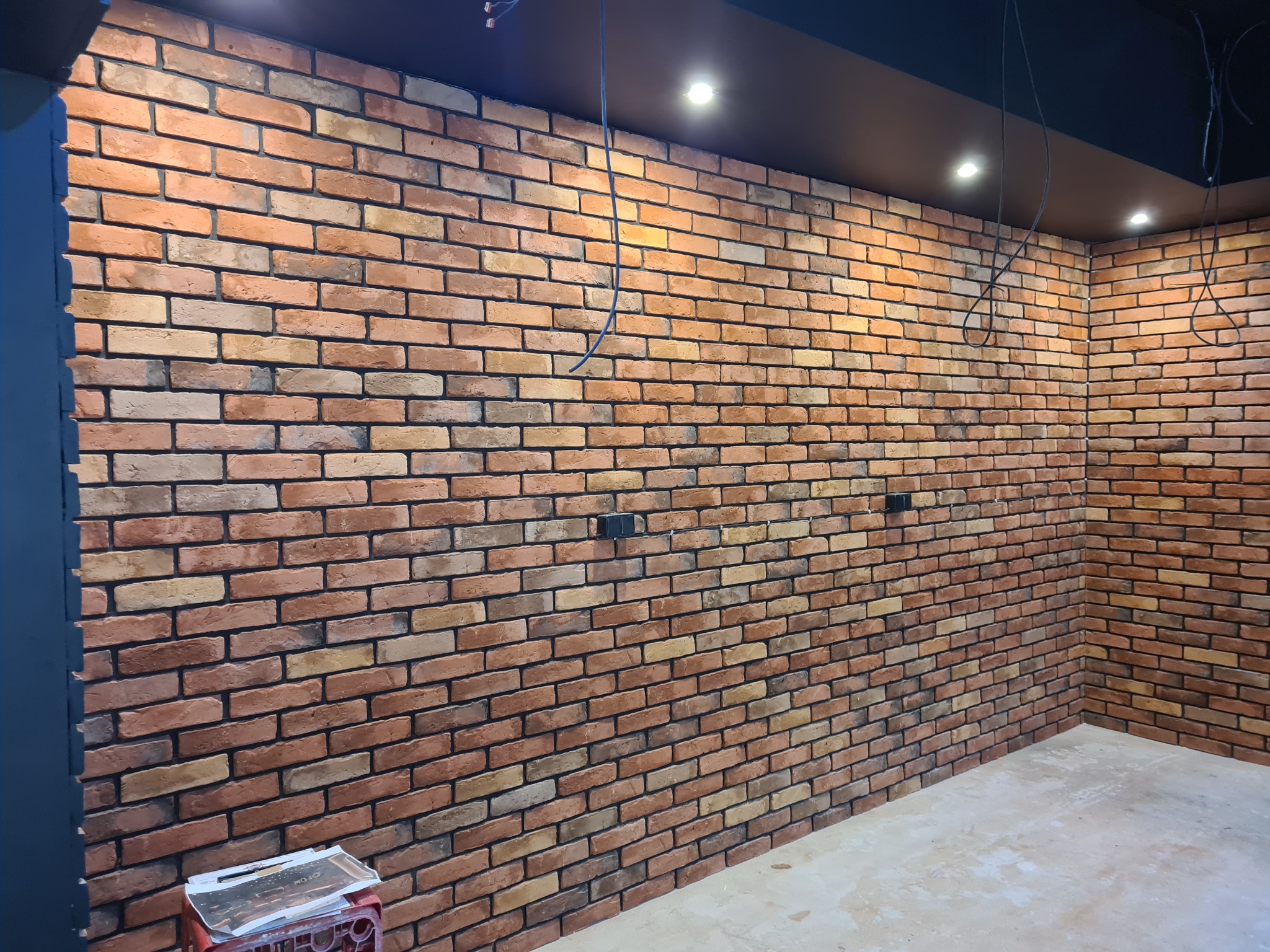 Ściana wykończona cegłą klinkierową w odcieniach czerwieni i beżu, widoczne niedokończone podłączenia elektryczne i surowa podłoga.