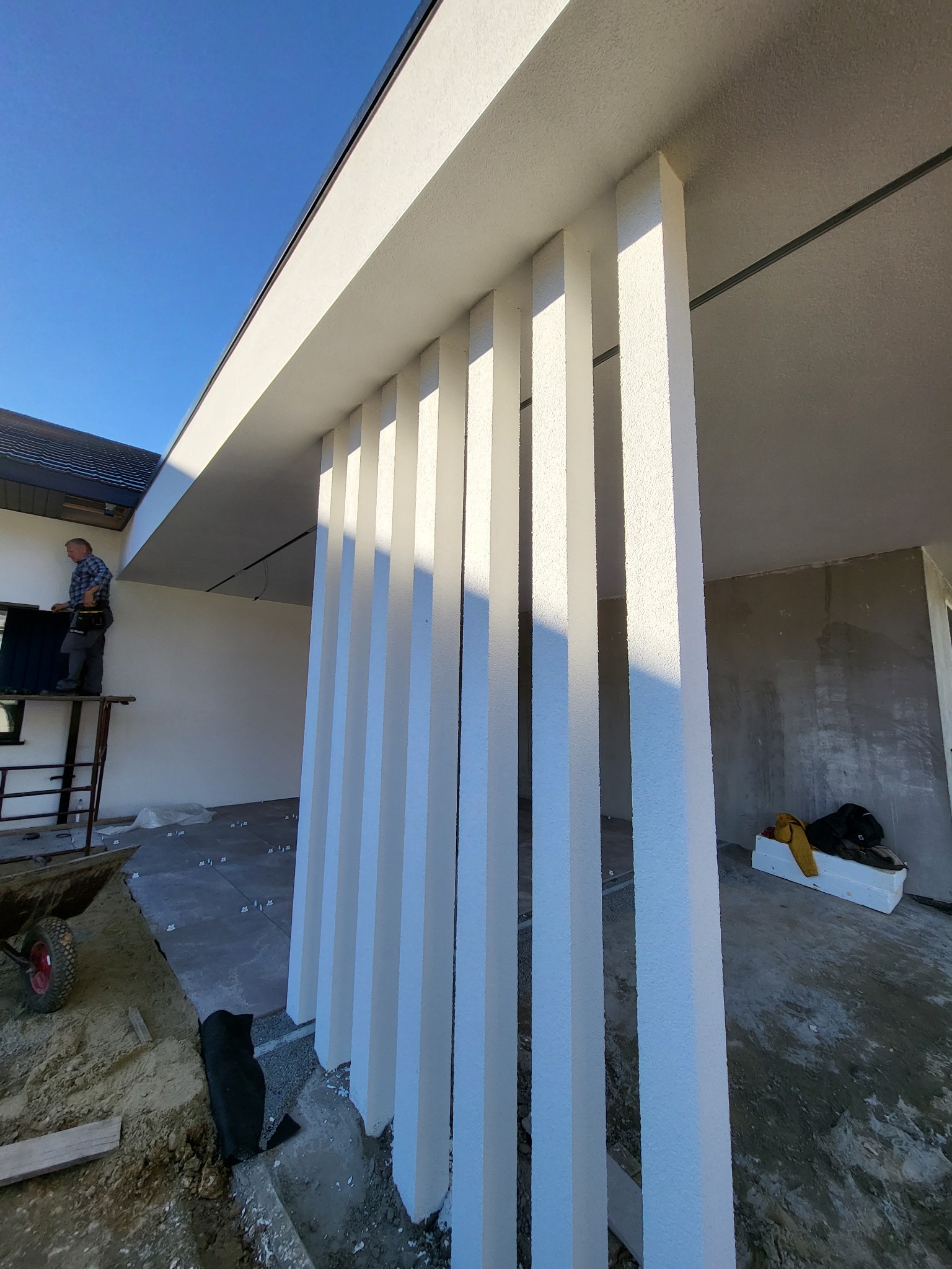 Budynek w trakcie prac wykończeniowych: widoczne białe, pionowe elementy dekoracyjne elewacji, pracownik na rusztowaniu montujący okno, surowy stan wnętrza, materiały budowlane na podłożu.