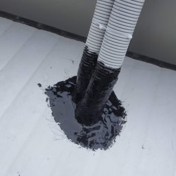 Kamil Matylek Klimat montaż serwis sprzedaż klimatyzacji i wentylacji - Dwa szare, karbowane przewody wychodzące z białej powierzchni, uszczelnione czarną substancją.