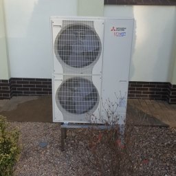 Kamil Matylek Klimat montaż serwis sprzedaż klimatyzacji i wentylacji - Biała, dwu-wentylatorowa pompa ciepła Mitsubishi Zubadan zamontowana na metalowej podstawie przed budynkiem z oknem witrażowym i kamiennym podłożem.