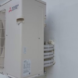 Kamil Matylek Klimat montaż serwis sprzedaż klimatyzacji i wentylacji - Zewnętrzna jednostka klimatyzacji Mitsubishi Electric zamontowana na białej ścianie, widoczne rury w izolacji przymocowane do ściany.