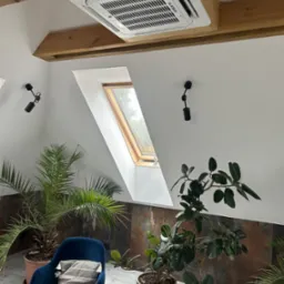 Kasetonowa jednostka klimatyzacyjna marki Kaisai zamontowana pod drewnianym sufitem w pomieszczeniu z oknem dachowym, oświetleniem punktowym i roślinami doniczkowymi.