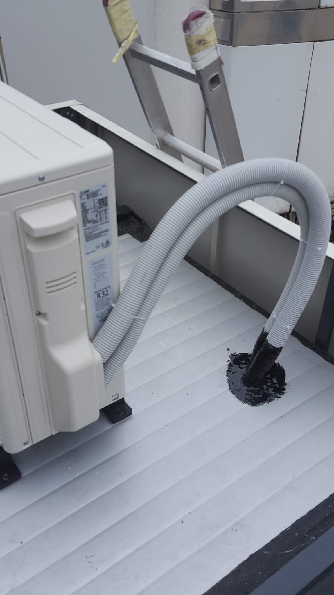 Zewnętrzna jednostka klimatyzatora marki Mitsubishi, z podwójnym przewodem odprowadzającym wilgoć, zamontowana na białym, żebrowanym dachu z widocznymi śladami uszczelniania wokół otworu...