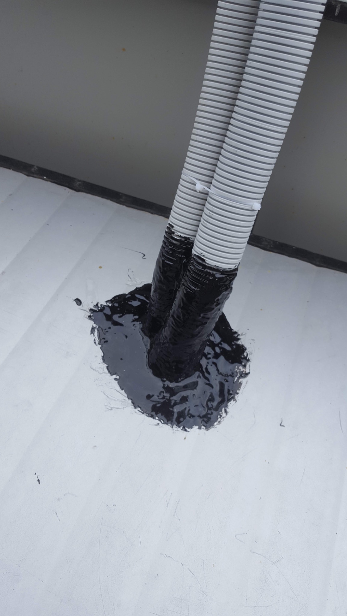 Dwa szare, karbowane przewody wychodzące z białej powierzchni, uszczelnione czarną substancją.