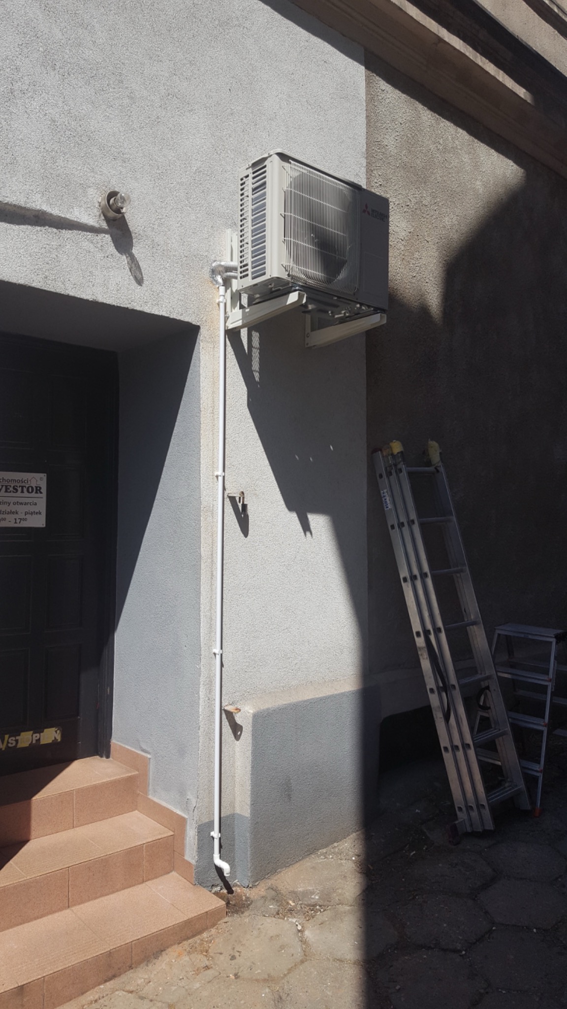 Zewnętrzna jednostka klimatyzacji Mitsubishi Electric zamontowana na szarej ścianie budynku obok ciemnych drzwi wejściowych i drabiny.