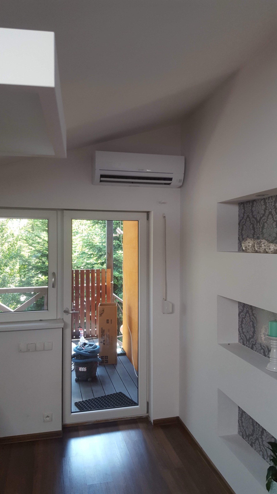 Biały klimatyzator ścienny zamontowany w nowoczesnym wnętrzu domu nad przeszklonymi drzwiami balkonowymi, obok dekoracyjnych wnęk ściennych i parkietu.