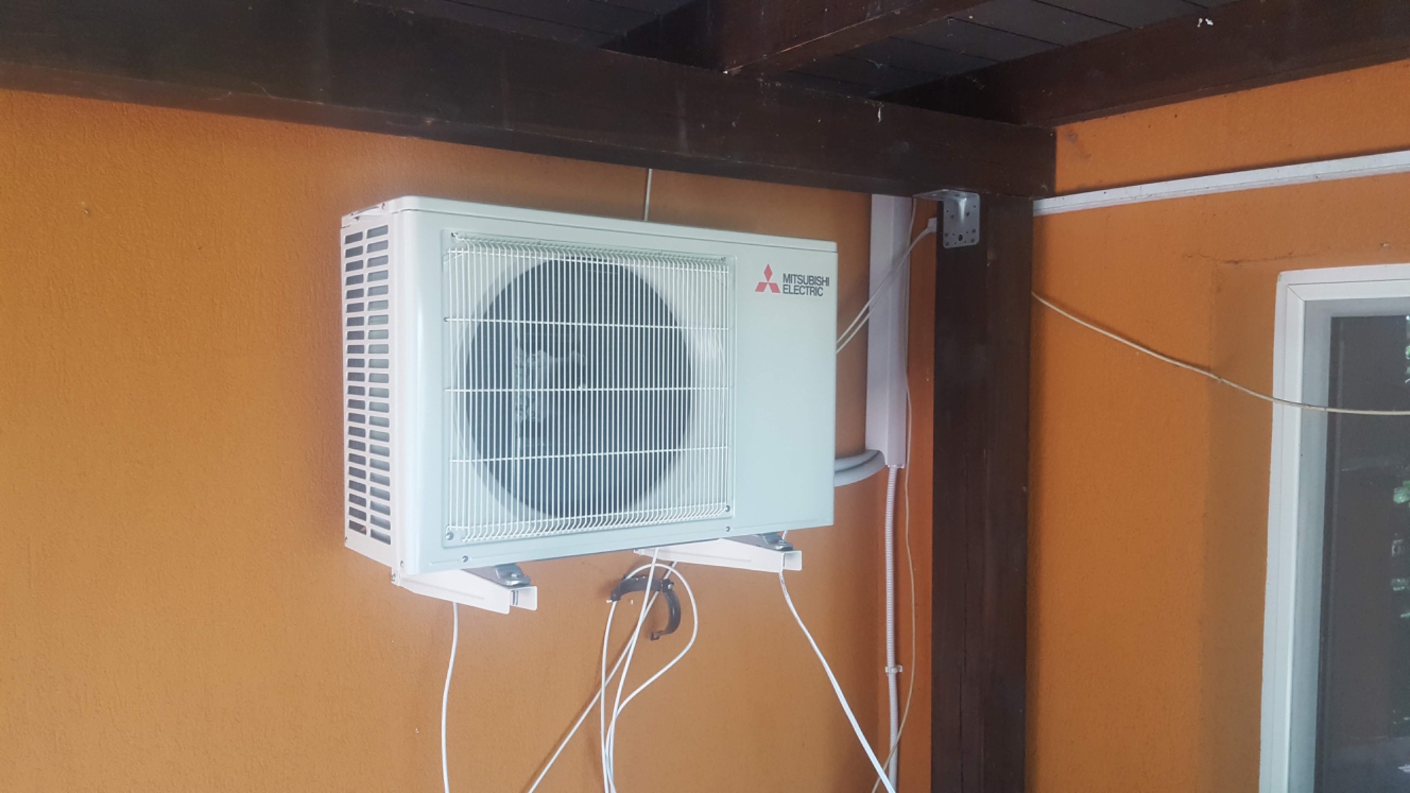 Zewnętrzna jednostka klimatyzatora Mitsubishi Electric zamontowana na pomarańczowej ścianie budynku z widocznymi przewodami i elementami instalacji.