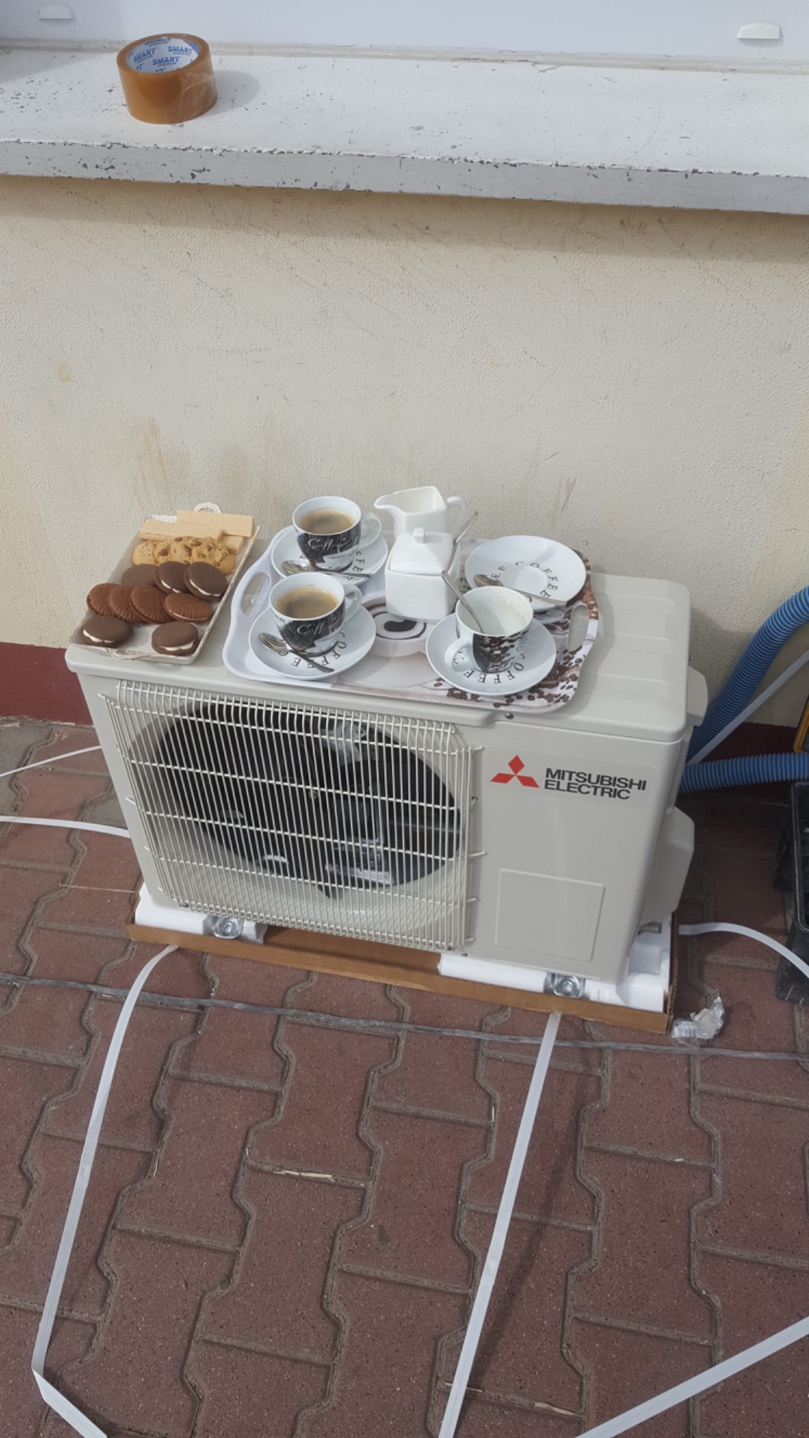 Zewnętrzna jednostka klimatyzacji Mitsubishi Electric z filiżankami kawy i ciasteczkami na górze, stojąca na palecie na tle ceglanej podłogi i beżowej ściany.