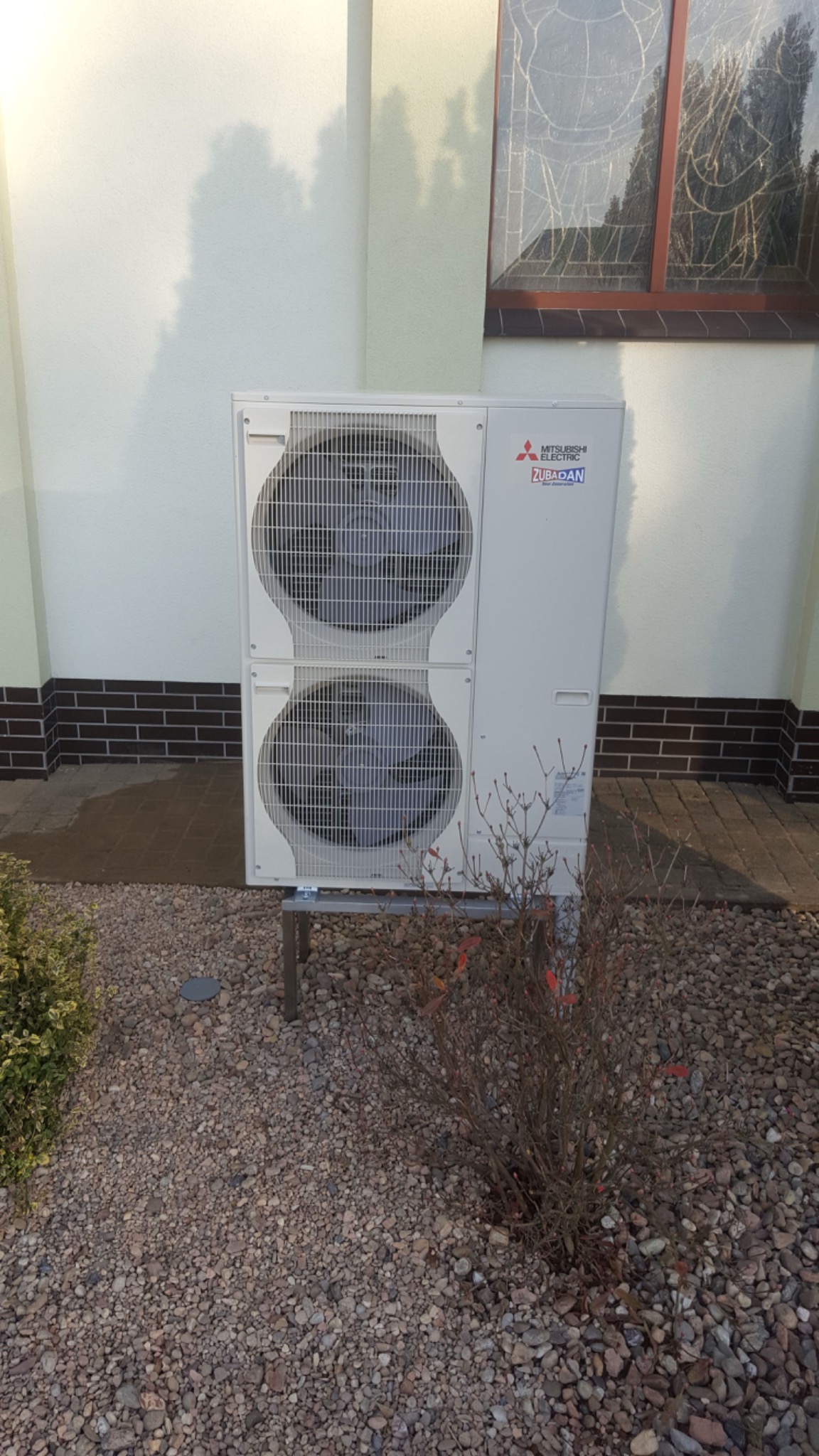 Biała, dwu-wentylatorowa pompa ciepła Mitsubishi Zubadan zamontowana na metalowej podstawie przed budynkiem z oknem witrażowym i kamiennym podłożem.