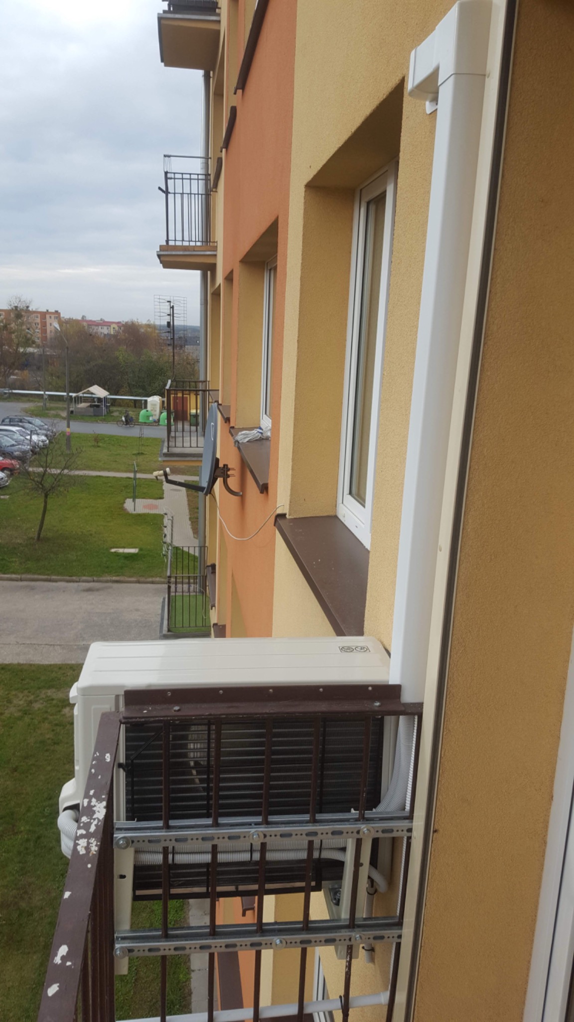 Zewnętrzna jednostka klimatyzacji zamontowana na balkonie bloku mieszkalnego, widok z boku, z brązową balustradą i elewacją w odcieniach pomarańczy i żółci.