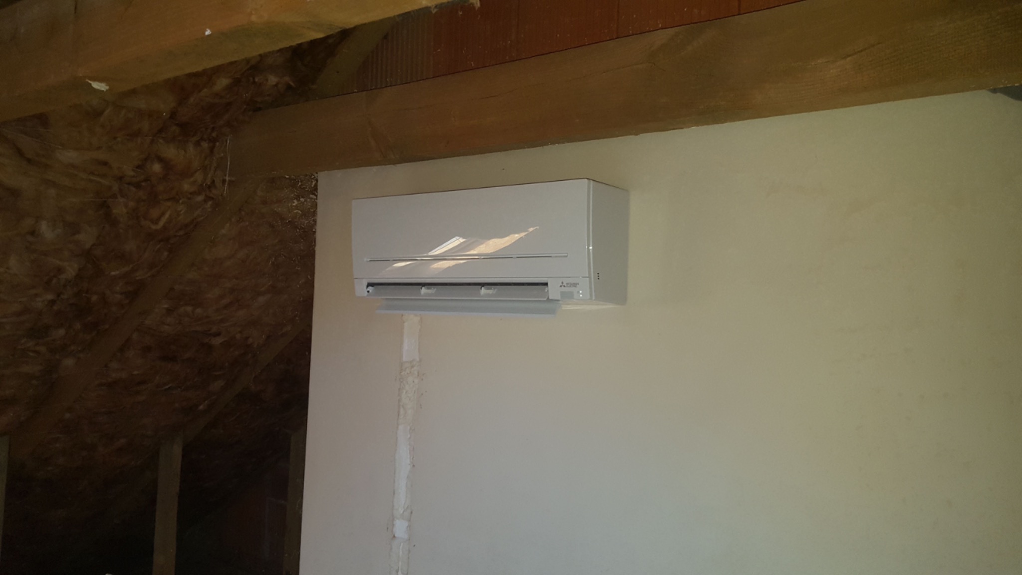 Biała jednostka klimatyzacyjna Mitsubishi zamontowana na ścianie w pomieszczeniu z widoczną drewnianą konstrukcją dachu.