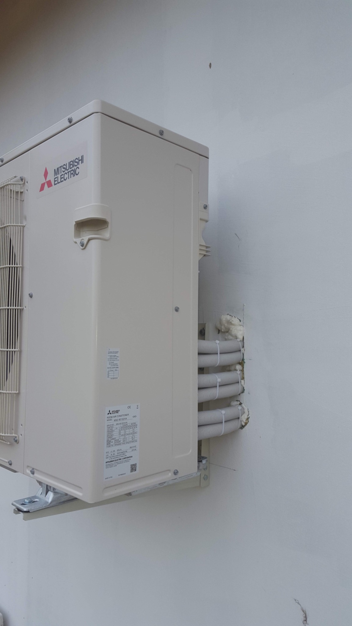 Zewnętrzna jednostka klimatyzacji Mitsubishi Electric zamontowana na białej ścianie, widoczne rury w izolacji przymocowane do ściany.