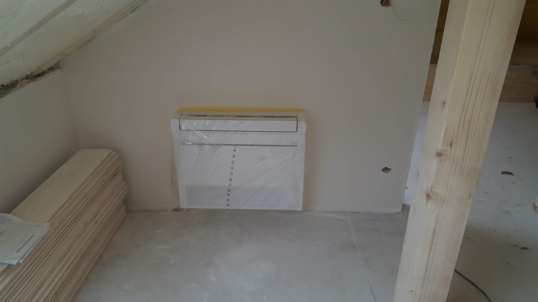 Nowa jednostka wewnętrzna klimatyzatora, zabezpieczona folią ochronną i taśmą, zamontowana na ścianie w surowym wnętrzu poddasza podczas remontu.