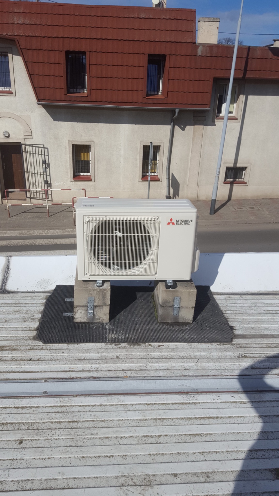 Zewnętrzna jednostka klimatyzacyjna Mitsubishi Electric zamontowana na betonowych blokach na dachu budynku, widok z niskiej perspektywy.