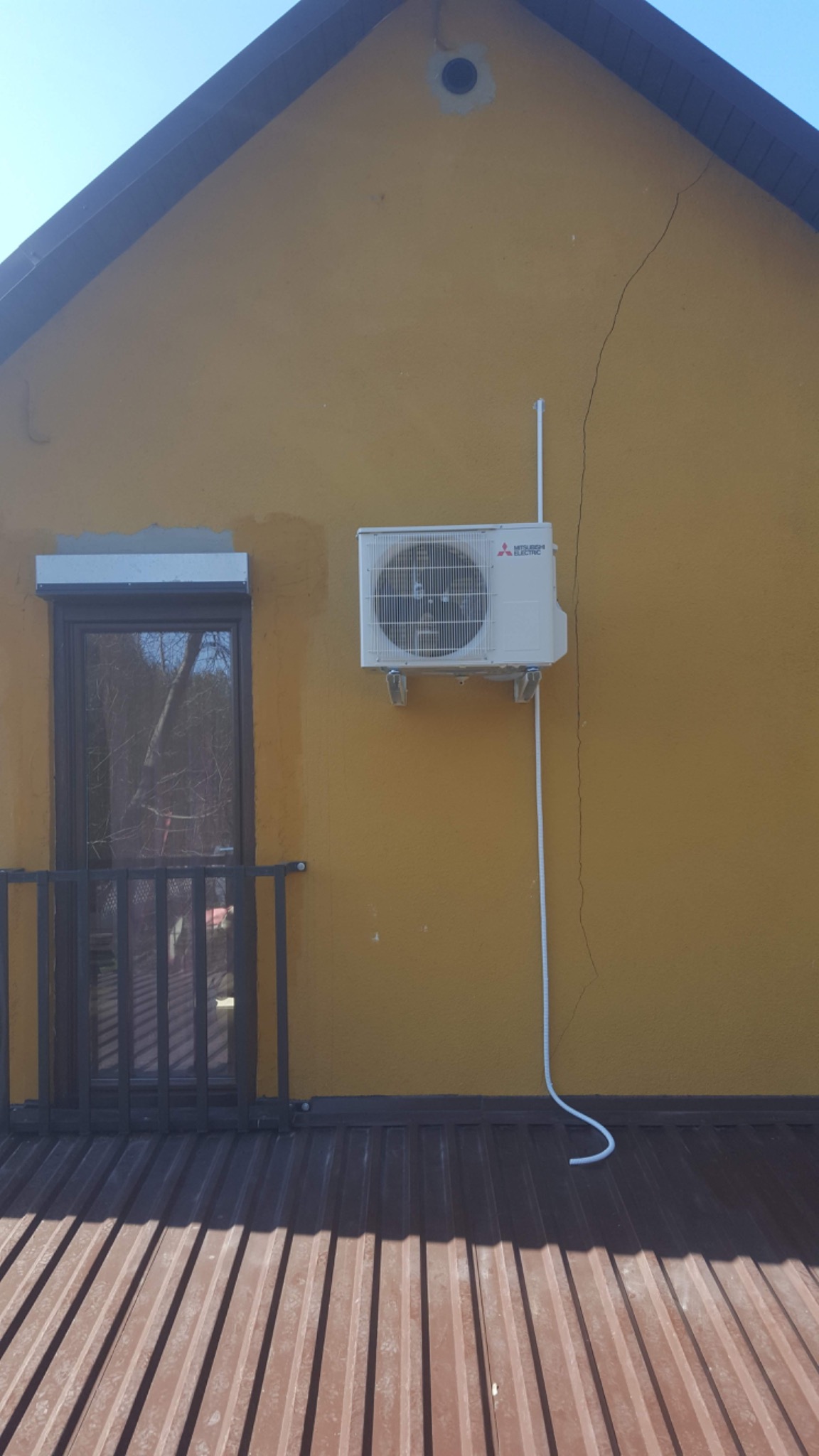 Zewnętrzna jednostka klimatyzatora Mitsubishi Electric zamontowana na żółtej ścianie domu, widoczna instalacja elektryczna i balkon z metalową barierką.