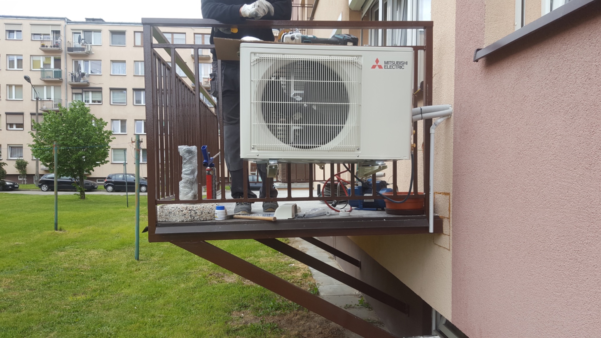 Montaż zewnętrznej jednostki klimatyzacji Mitsubishi Electric na balkonie, widoczne narzędzia i osprzęt instalacyjny, w tle budynek mieszkalny.