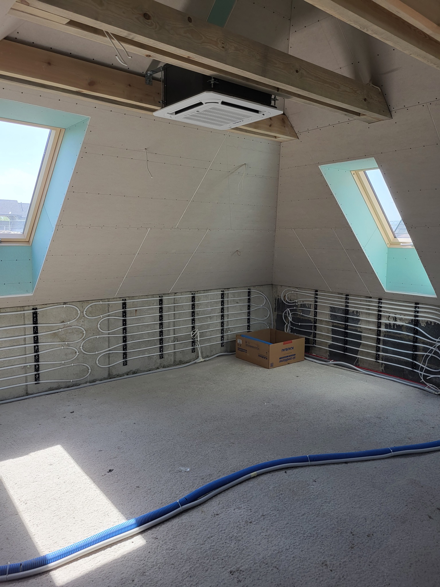 Wnętrze poddasza w trakcie instalacji: widoczny zamontowany klimatyzator kasetonowy, okna dachowe, instalacja ogrzewania podłogowego na ścianach oraz kartonowe pudełko po klimatyzatorze.