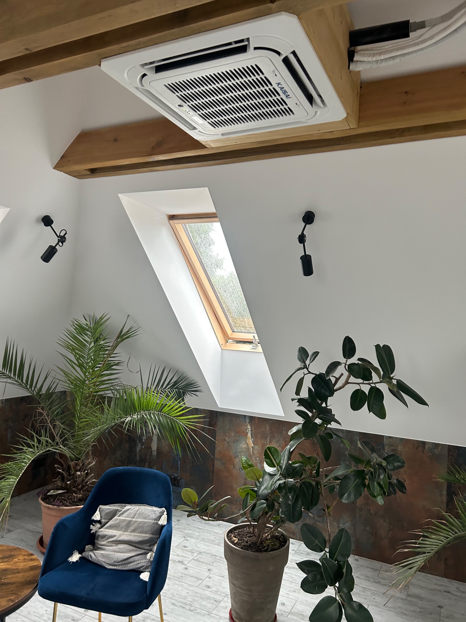 Kasetonowa jednostka klimatyzacyjna marki Kaisai zamontowana pod drewnianym sufitem w pomieszczeniu z oknem dachowym, oświetleniem punktowym i roślinami doniczkowymi.
