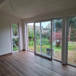 RAFAŁ PYC - Jasne wnętrze z panelami podłogowymi i dużymi oknami z widokiem na ogród. Widoczny odkurzacz przemysłowy przy przesuwanych drzwiach balkonowych.