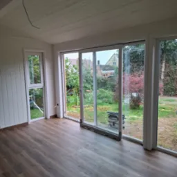 Jasne wnętrze z panelami podłogowymi i dużymi oknami z widokiem na ogród. Widoczny odkurzacz przemysłowy przy przesuwanych drzwiach balkonowych.