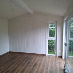 RAFAŁ PYC - Wnętrze domku szkieletowego z drewnianą podłogą, białymi panelami ściennymi i dużym oknem z widokiem na zieleń. Widoczny kabel zwisający z sufitu.