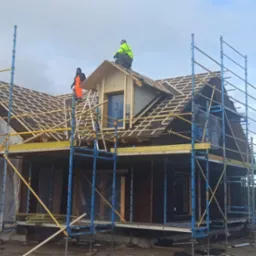 Budowa domu z ciemnego drewna, widoczne rusztowania, więźba dachowa w trakcie montażu, dwaj pracownicy w odblaskowych ubraniach na dachu, pochmurne niebo w tle.