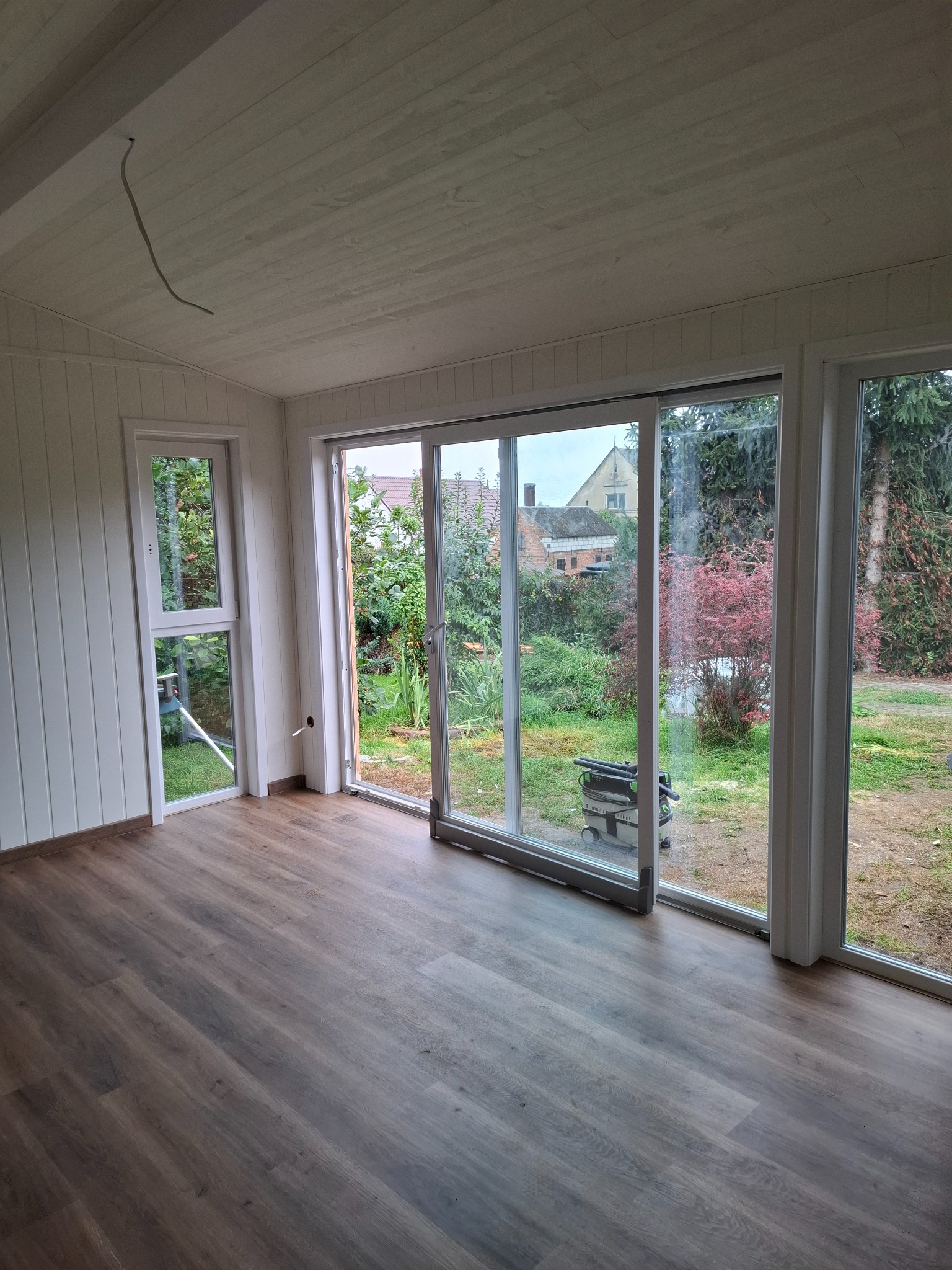 Jasne wnętrze z panelami podłogowymi i dużymi oknami z widokiem na ogród. Widoczny odkurzacz przemysłowy przy przesuwanych drzwiach balkonowych.