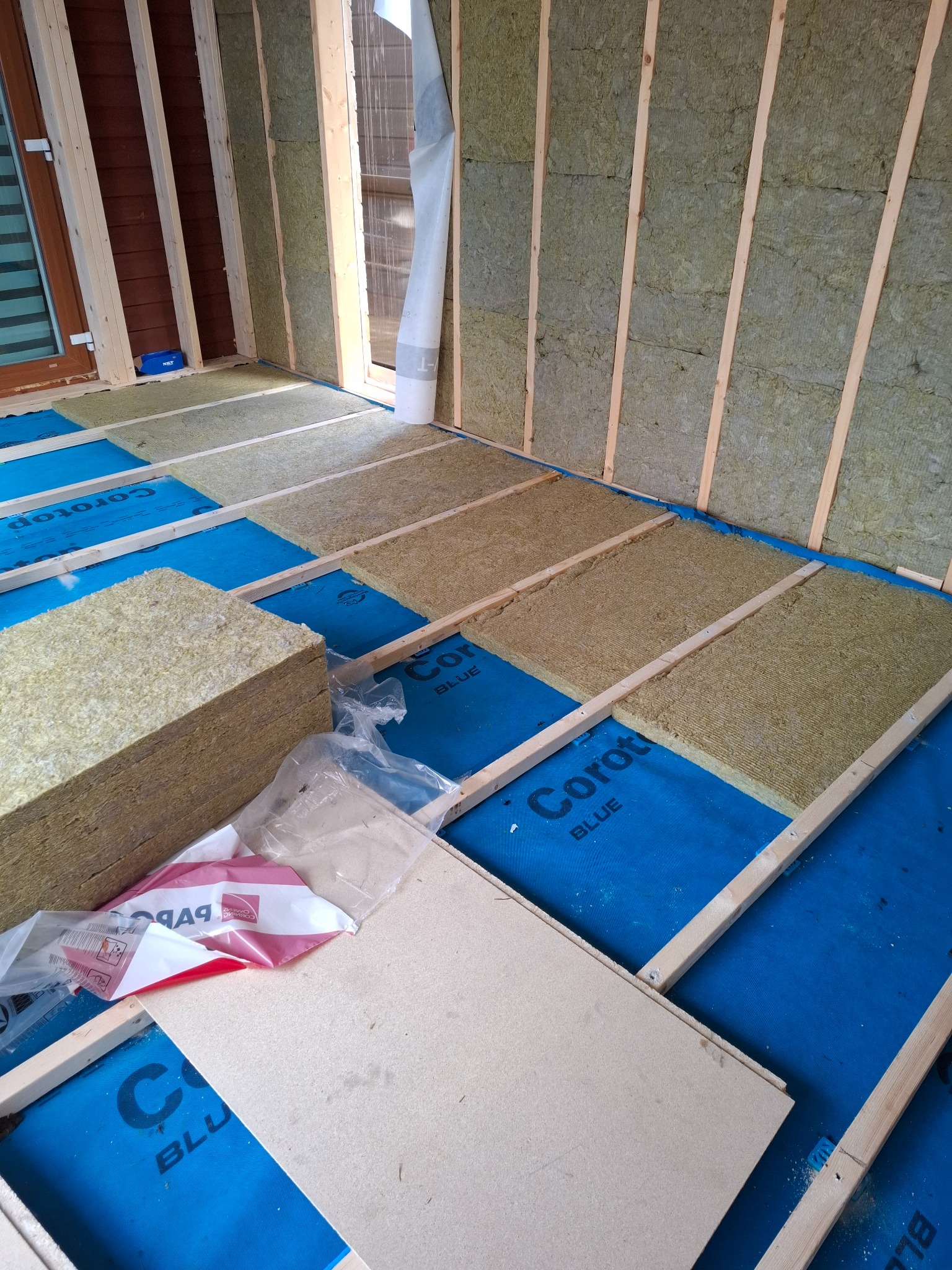 Ocieplenie ścian i podłogi budynku w trakcie budowy. Widoczne drewniane legary, izolacja z wełny mineralnej i folia paroizolacyjna w kolorze niebieskim.