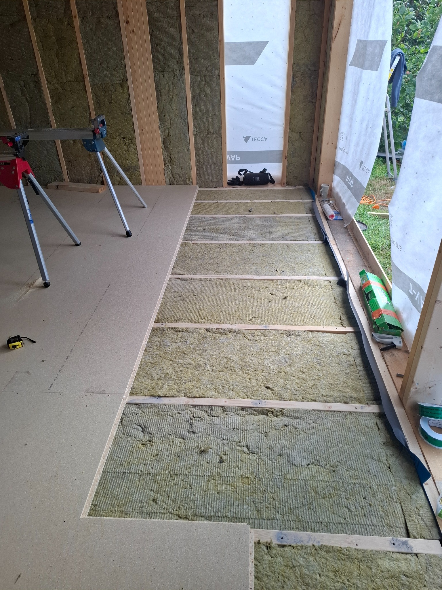 Wnętrze w trakcie budowy: izolacja ścian wełną mineralną, częściowo ułożone panele podłogowe na legarach. Widoczne narzędzia i materiały budowlane.