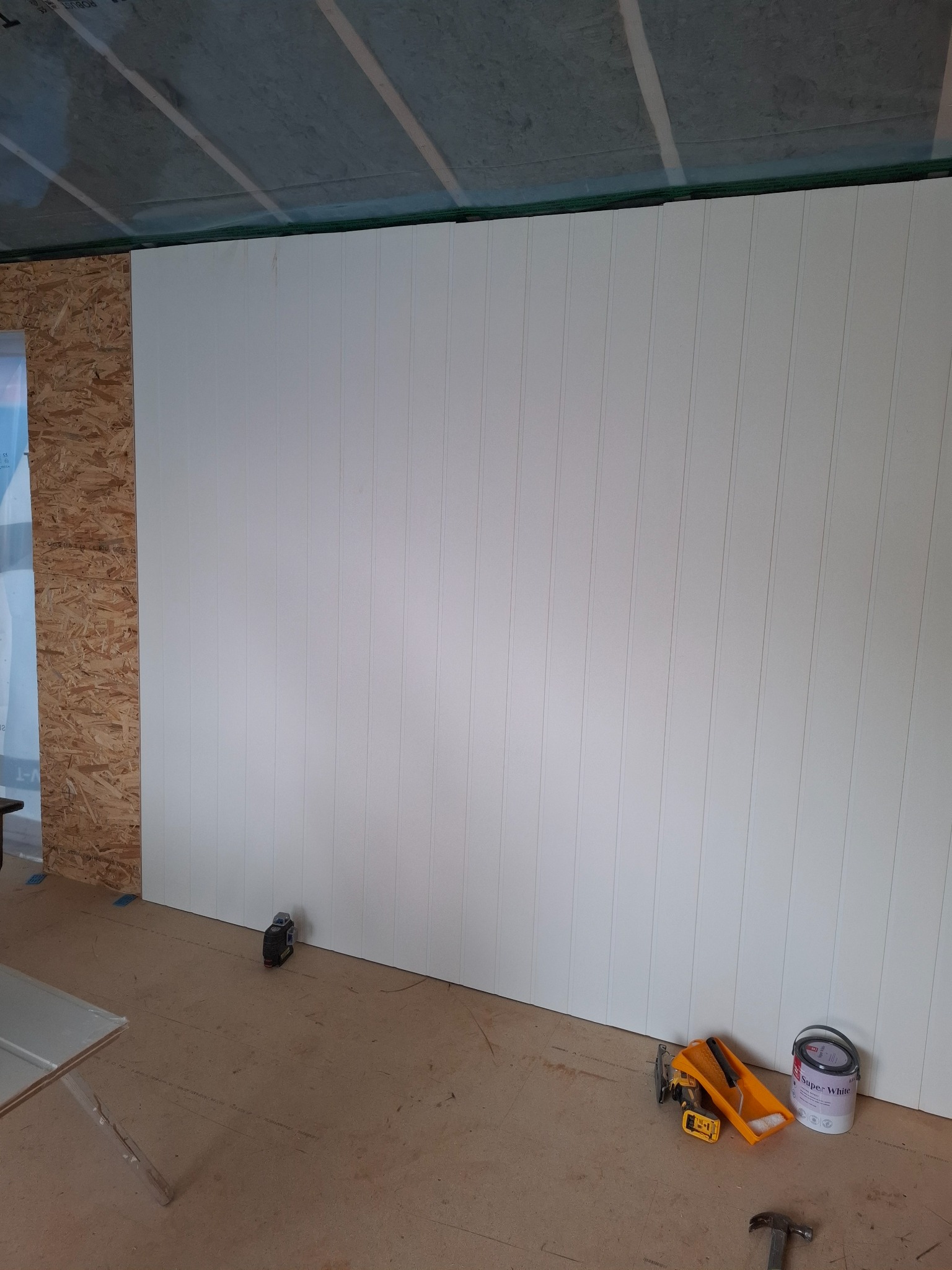 Wnętrze w trakcie wykańczania: białe panele ścienne, surowa podłoga, narzędzia i puszka farby. Częściowo wykończona ściana z płyty OSB z lewej strony.