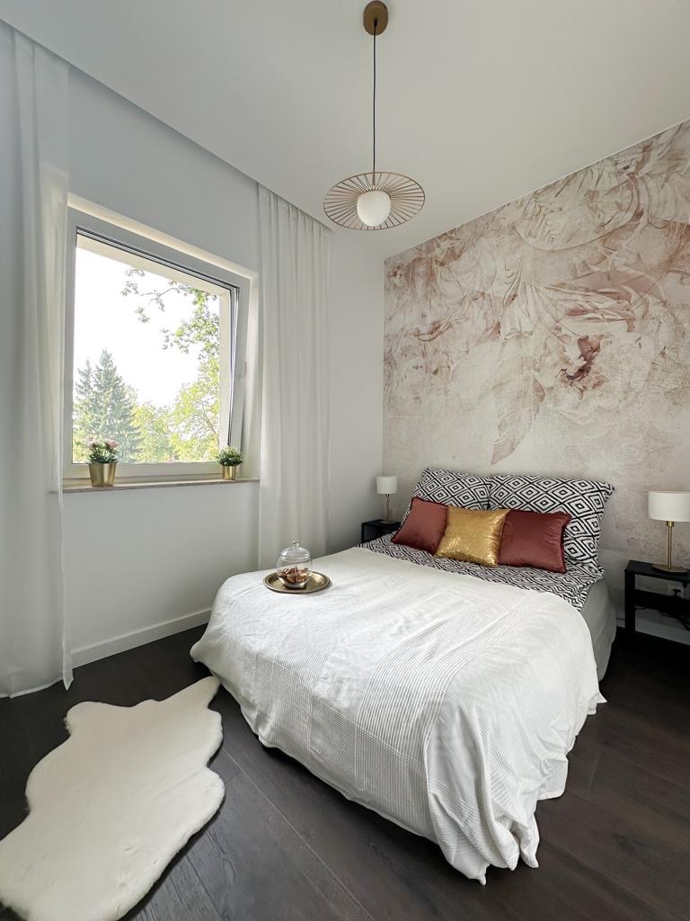 Jasna sypialnia z łóżkiem, dekoracyjną ścianą i lampą wiszącą, uzupełniona o biały dywanik i stoliki nocne z lampkami.
