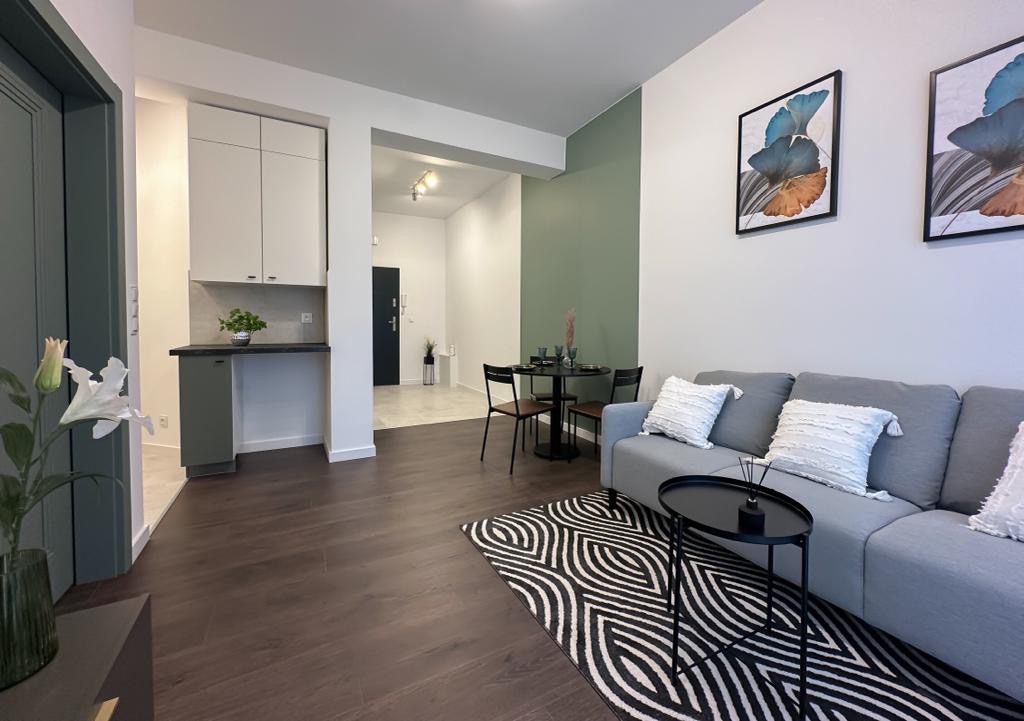 Jasne wnętrze mieszkania po remoncie: ciemna drewniana podłoga, szara sofa z dekoracyjnymi poduszkami, okrągły stolik kawowy, czarno-biały dywan w geometryczny wzór, aneks kuchenny z zielonymi...