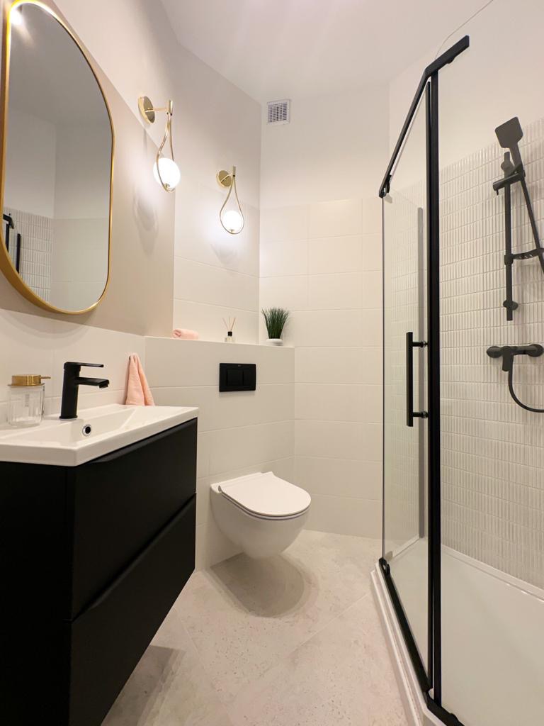 Nowoczesna łazienka w bieli i czerni z akcentami złota: czarna szafka pod umywalką, owalne lustro w złotej ramie, minimalistyczna kabina prysznicowa z czarnymi profilami, wisząca toaleta i złote...