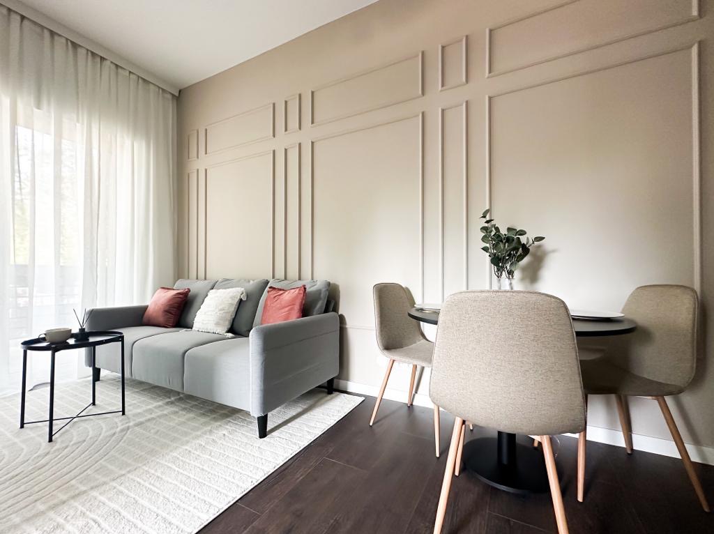 Jasne wnętrze mieszkania z szarą sofą z poduszkami, okrągłym stolikiem kawowym, dywanem w geometryczne wzory, stołem z krzesłami i dekoracyjną ścianą z listwami.