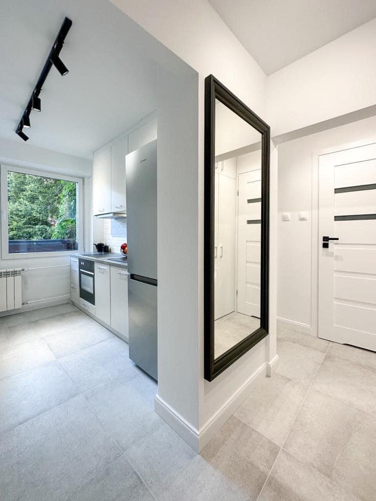 Nowoczesne wnętrze: jasna kuchnia z białymi szafkami, srebrną lodówką i szarymi płytkami podłogowymi, widok na przedpokój z dużym lustrem w czarnej ramie i białymi drzwiami z czarnymi akcentami.