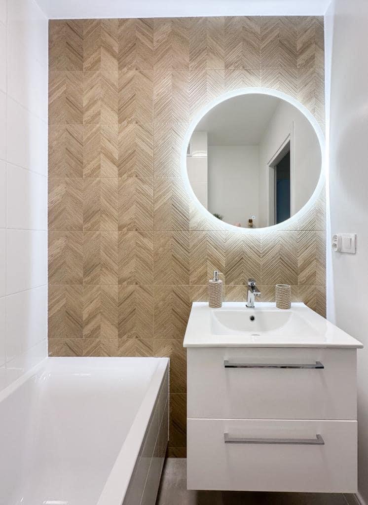 Nowoczesna łazienka z umywalką w białej szafce z dwoma szufladami, okrągłym lustrem podświetlanym LED i ścianą wyłożoną płytkami w jodełkę imitującymi drewno.