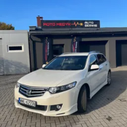 Białe kombi Honda Accord z pakietem stylistycznym stoi przed budynkiem warsztatu Moto Medyk Auto Serwis z czarnym szyldem i flagami reklamowymi.