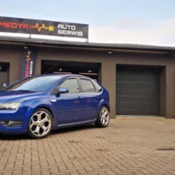 Niebieski Ford Focus zaparkowany przed warsztatem samochodowym Moto Medyk Auto Serwis, widoczny szyld i flaga reklamowa, w tle brama garażowa oraz baner ProfiAutoSerwis.
